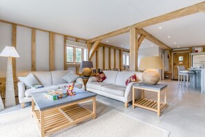 A spacious living room with a high ceiling, exposed wooden beams, a sofa set, a coffee table, and a kitchen area with a washing machine.