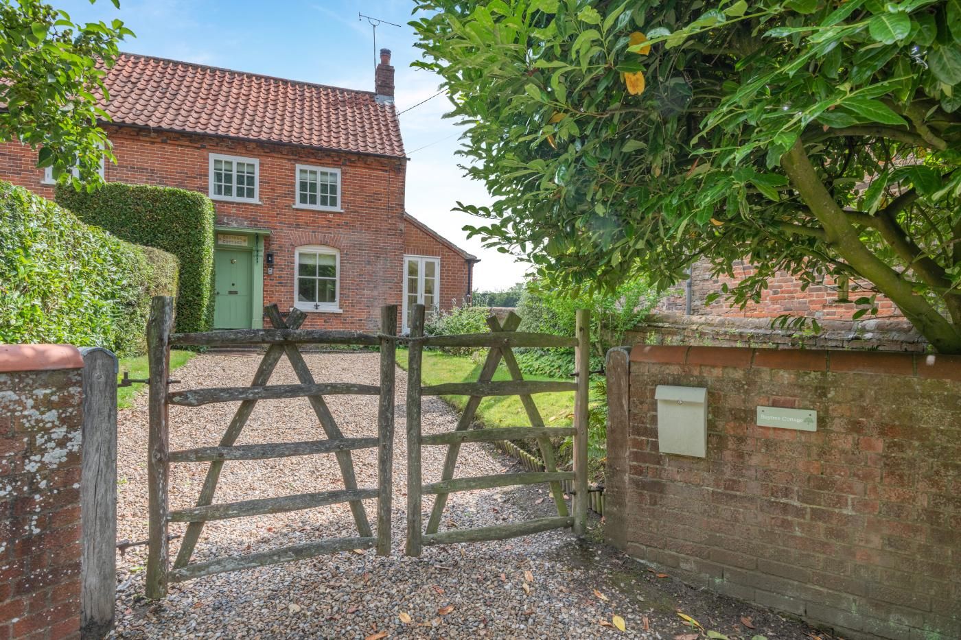 A brick house with a red-tiled roof, a green door, and a gravel driveway leading to a wooden gate.