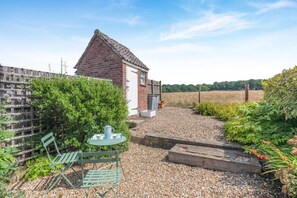 A rustic house with a brick exterior, a small garden area with a table and chairs, and a gravel path leading to a wooden deck. - 1 Bed in Swanton Novers (oc-1747) (Swanton Novers)