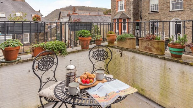 A balcony with a table set for breakfast, surrounded by potted plants and a view of a stone building. - 2 Bed in Lynton (oc-s33456) (Lynmouth)