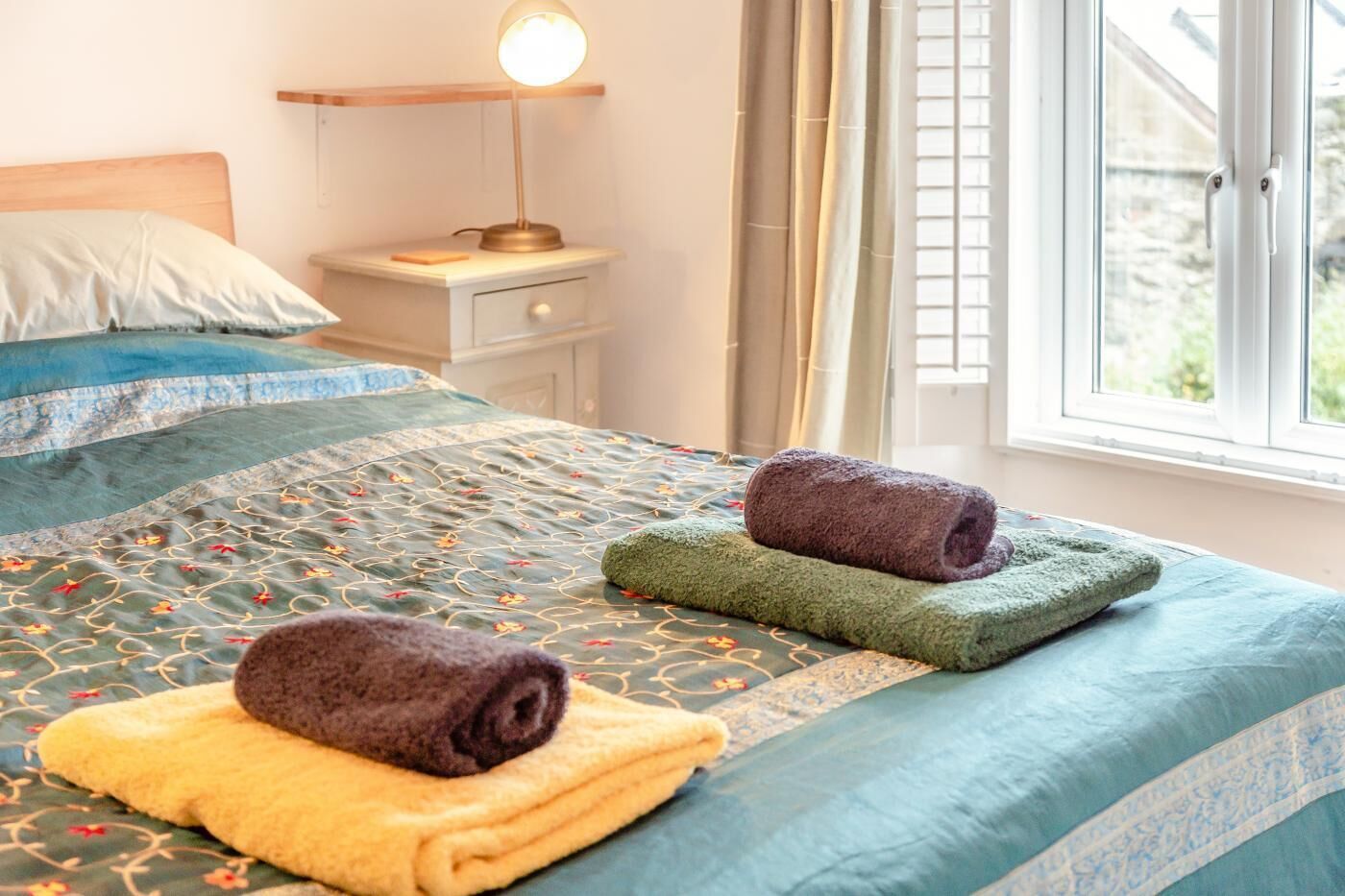 A neatly made bed with a patterned bedspread, two rolled towels, a bedside lamp, and a window with curtains.