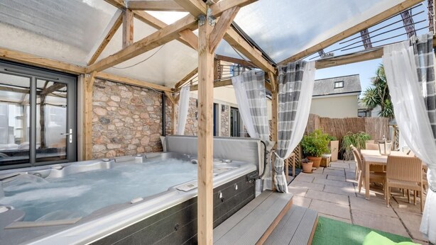 Et område med jacuzzi, stenvægge og træbjælker, en overdækket terrasse med fletmøbler og udsigt over det omkringliggende kvarter.