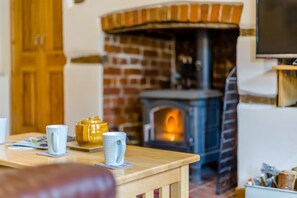 A cozy living room with a brick fireplace, a wooden table with mugs, and a television on a stand. - 2 Bed in Reepham (oc-1755) (Reepham)