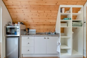 A compact kitchen with a microwave, refrigerator, and white cabinetry. A wooden ceiling and a white shelving unit with folded towels are also visible.