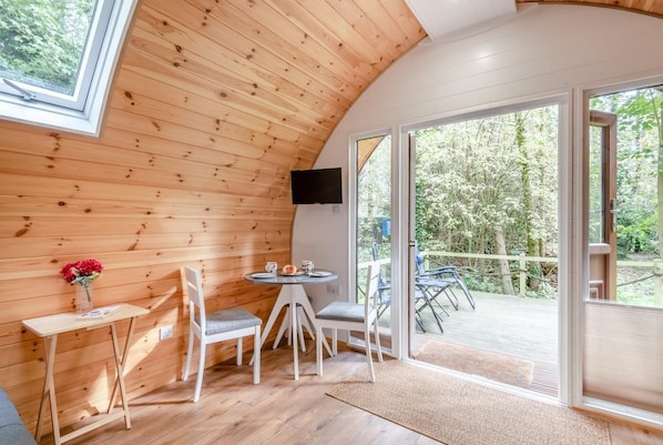 A cozy cabin with wooden walls, a round table with chairs, a TV mounted on the wall, and a large sliding glass door leading to an outdoor deck.