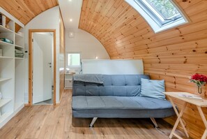 A cozy room with a curved wooden ceiling, a grey sofa, a small table with a vase of flowers, and a window with a view of greenery.