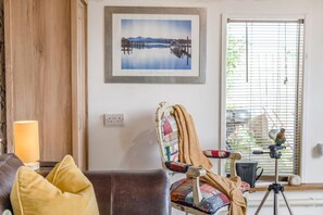 A living room with a wooden wall, a framed photograph, a lamp, a chair with a blanket, and a tripod with a camera. - 1 Bed in Totnes (oc-bc1000) (Totnes)