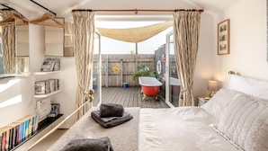 Ein Schlafzimmer mit Bett, Bücherregal, einem Fenster mit Vorhängen, einer roten Badewanne und Blick ins Freie.