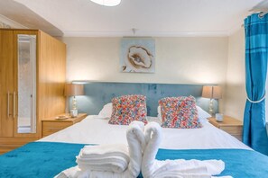 A hotel room with a large bed, two pillows with a floral pattern, a blue headboard, a wooden wardrobe, and a painting on the wall.