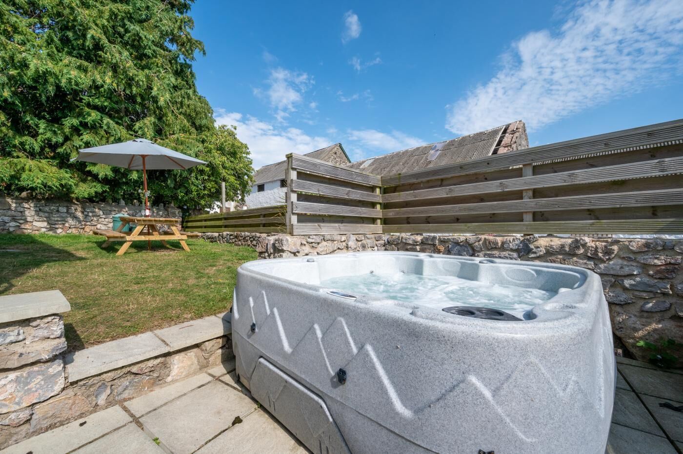 A hot tub in a backyard with stone walls, a picnic table, and a wooden deck.