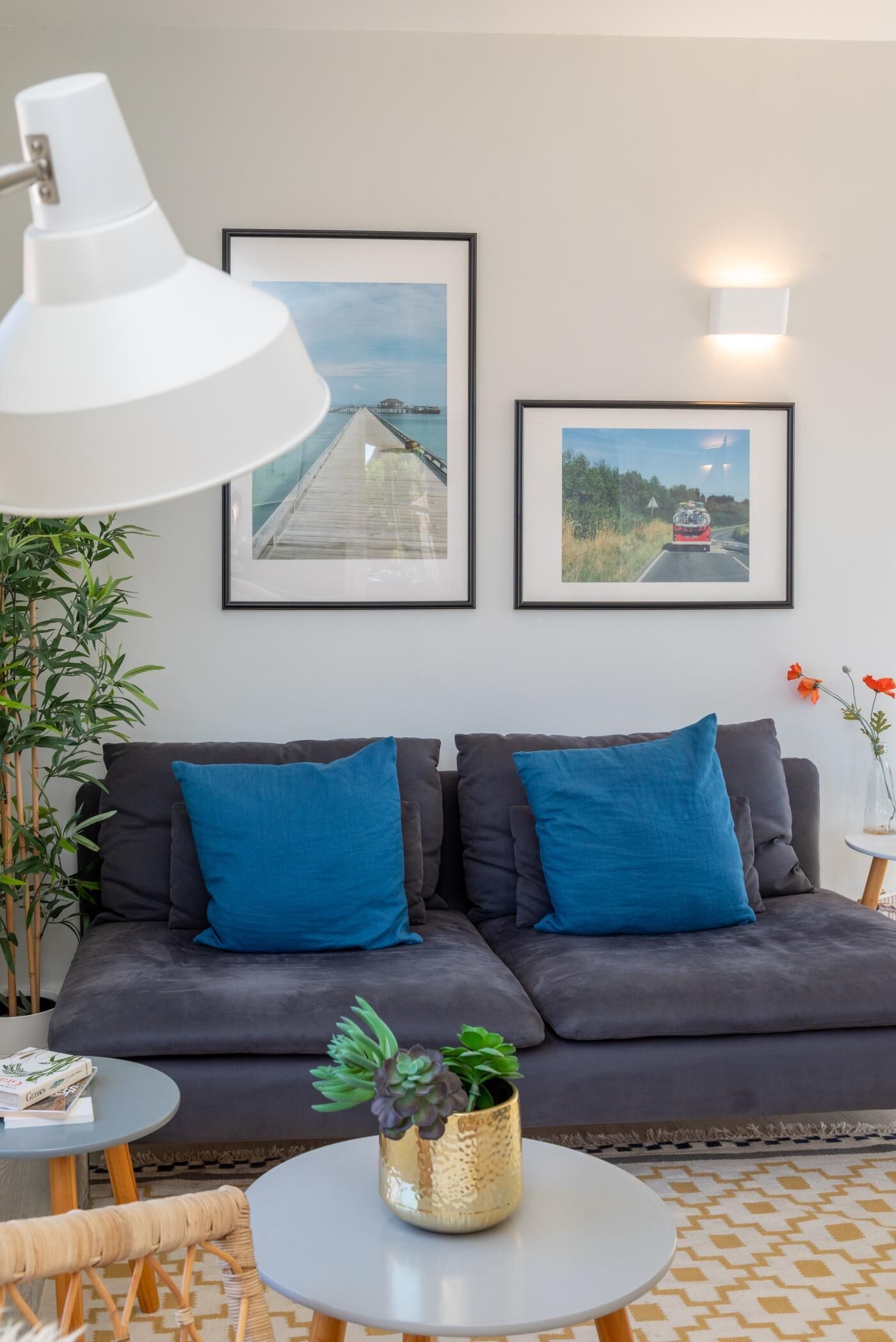 Un salon moderne avec un canapé gris foncé, deux coussins bleus, une lampe de bureau blanche, deux tableaux encadrés accrochés au mur, une petite table ronde avec une plante et un tapis à motifs.