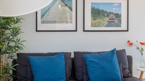 Ein modernes Wohnzimmer mit einem dunkelgrauen Sofa, zwei blauen Kissen, einer weißen Schreibtischlampe, zwei gerahmten Bildern an der Wand, einem kleinen runden Tisch mit einer Pflanze und einem gemusterten Teppich.