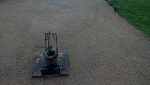 Un camino de grava que conduce a un edificio de piedra con una estatua, un estanque y un campo cubierto de césped.