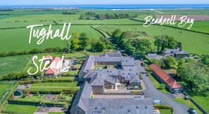 Vue aérienne de Tughall Steads, avec en premier plan un vaste domaine comprenant plusieurs bâtiments, une pelouse bien entretenue et une vue sur la baie de Beadnell.