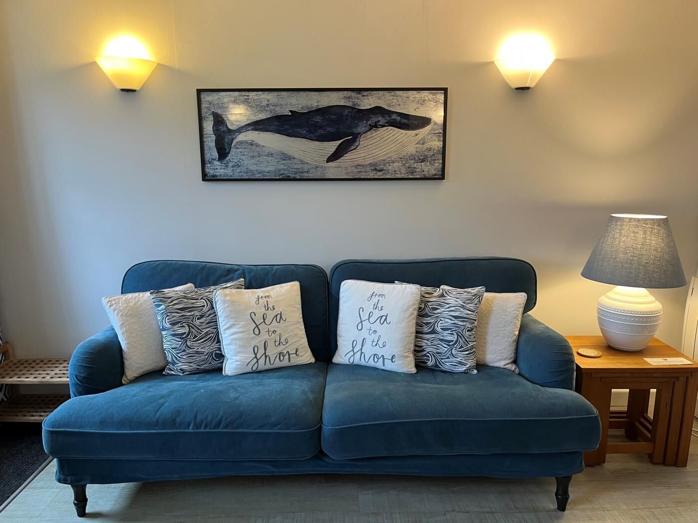 A blue sofa with four decorative pillows, a wall-mounted whale painting, and two wall sconces.