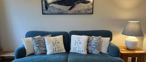 A blue sofa with four decorative pillows, a wall-mounted whale painting, and two wall sconces.