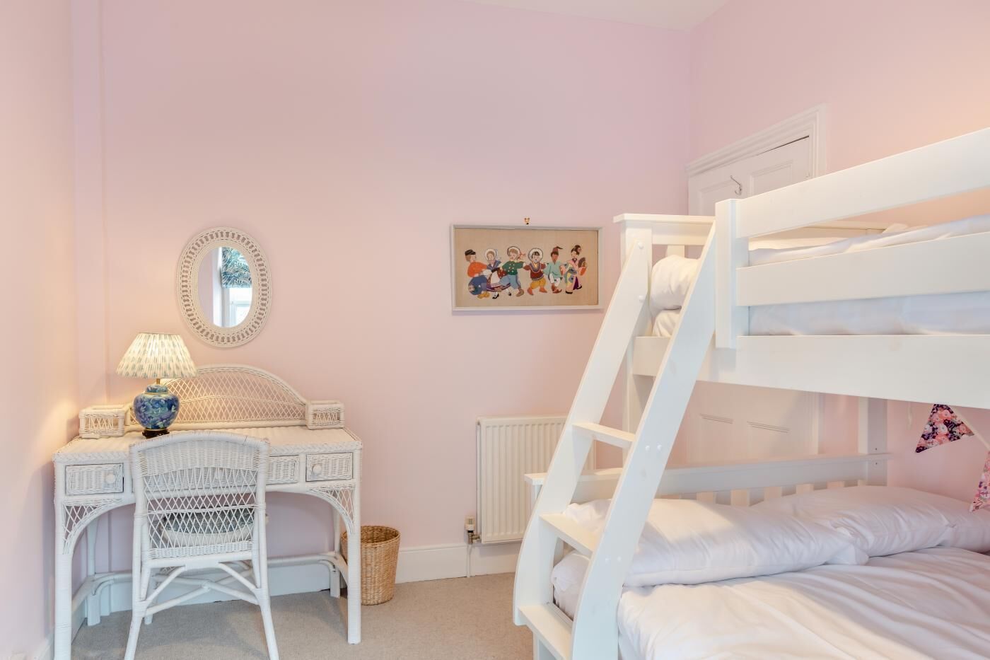 Une chambre avec un lit superposé, un bureau blanc avec une lampe, une chaise en osier et un tableau encadré accroché au mur.