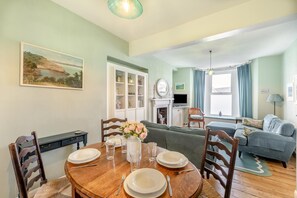 A dining area with a round wooden table set for four, a sofa, and a television in the background. - 4 Bed in Brixham (oc-b33461) (Brixham)