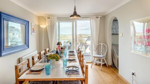 Comedor con una gran mesa dispuesta para cuatro, vista al mar a través de amplios ventanales, y una silla blanca con cojín.