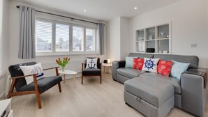 A modern living room with a grey sofa, a small table, and a chair. There is a window with blinds, a radiator, and a shelf with decorative items. - 2 Bed in Swanage (oc-wy606) (Swanage)