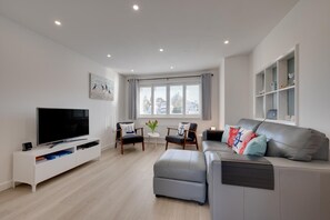 Un salon moderne avec un téléviseur à écran plat, un canapé gris, une petite table avec un vase et un fauteuil avec un coussin.