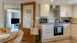 A compact kitchen with a microwave, oven, and a coffee maker. There's a dining area with a table and chairs, and a TV in the background. - 3 Bed in Petrockstow (oc-n33541) (Petrockstow)