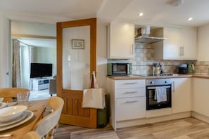 Cocina compacta con microondas, horno y cafetera. Hay un comedor con mesa y sillas, y un televisor al fondo.