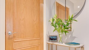 Een houten deur, een ronde spiegel, een witte console tafel met een vaas groene planten, en een klein lijstje met een foto.