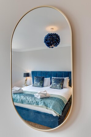 Une chambre à coucher avec un grand miroir ovale, une tête de lit recouverte d’un tissu bleu, un lit avec du linge de maison blanc et une décoration murale.