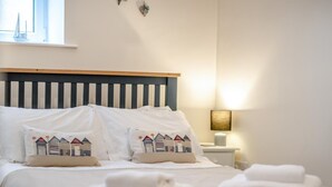 A neatly made bed with white linens and decorative pillows, a bedside table with a lamp, and a window with a view. - 2 Bed in Whitby (oc-w402) (Whitby)