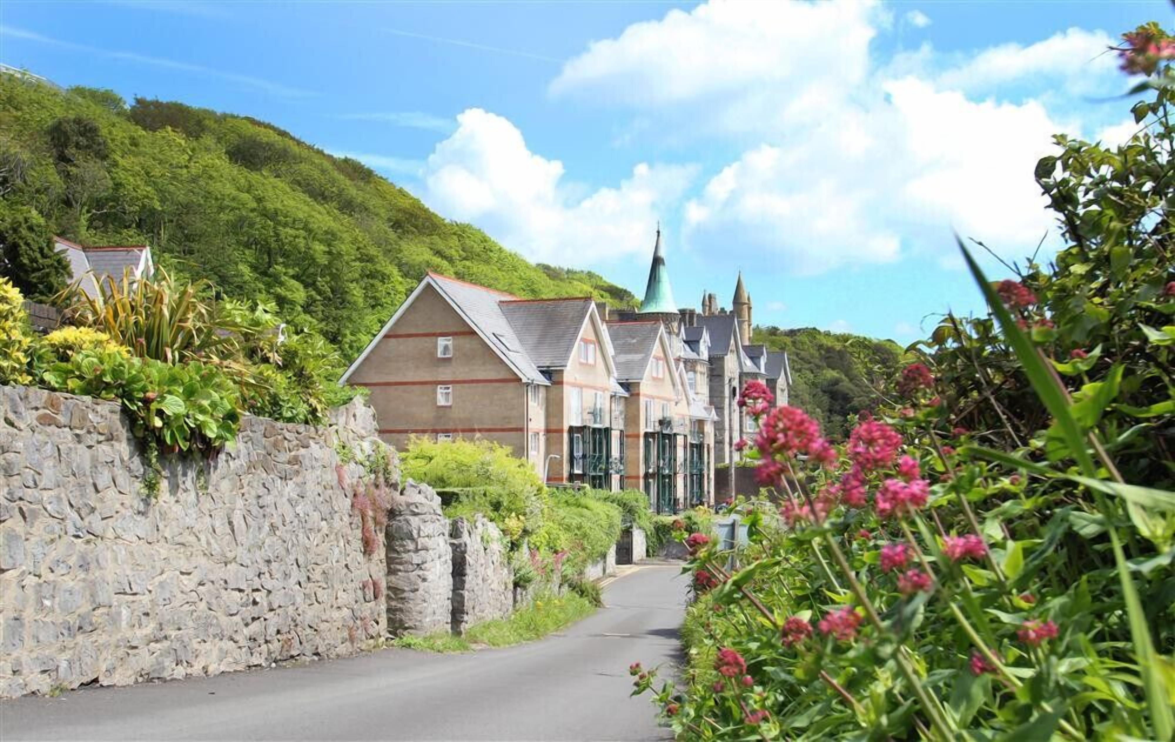 A narrow road flanked by stone walls and lush greenery, leading to a row of traditional buildings with steep roofs and a prominent church steeple in the background.