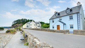 Eine Küstenstraße mit Steinmauern, Bänken und Häusern mit hellblau-weißen Fassaden.