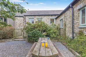 A stone building with a gravel courtyard, a wooden table with a vase of flowers, and a wooden fence.