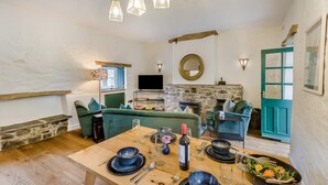 Ein gemütliches Wohnzimmer mit einem Kamin aus Stein, grünen Sofas, einem Holzesstisch für vier Personen und einem Fernseher.