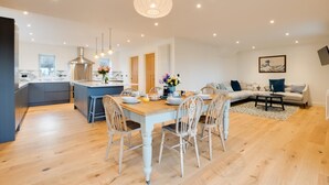 A modern kitchen with a central island, a dining area with a wooden table and chairs, and a living room with a sofa and a coffee table. - 4 Bed in Rock (oc-nb1952) (Rock)