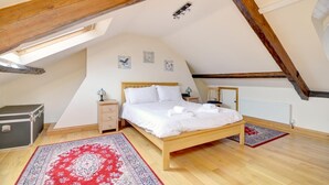 Ein Schlafzimmer mit schrägen Wänden, Holzbalken, einem Bett mit weißer Bettwäsche, zwei Nachttischen, einer Kommode, einem Heizkörper und zwei gemusterten Teppichen.