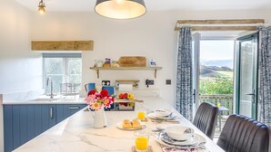 A modern kitchen with a marble countertop, a dining area with a table set for breakfast, and a view of the countryside through large windows.