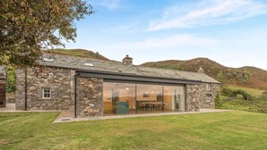 Une maison en pierre avec une extension en verre, une pelouse verdoyante et des collines en arrière-plan.