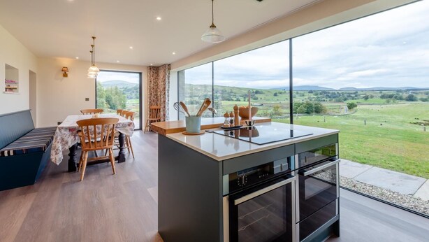 Une cuisine moderne dotée d’un grand îlot central, d’un coin repas avec une table et des chaises, et offrant une vue sur la campagne.