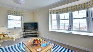 A living room with a television, a sofa, a coffee table with fruits and a book, and a view of the beach through large windows. - 5 Bed in Amroth (oc-pw9068) (Amroth)