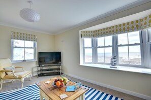 A living room with a television, a sofa, a coffee table with fruits and a book, and a view of the beach through large windows. - 5 Bed in Amroth (oc-pw9068) (Amroth)