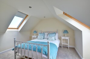 Ein Schlafzimmer mit schrägen Wänden, einem Oberlicht, einem Bett mit weißem Metallgestell, zwei Nachttischen mit Lampen und einem Teppichboden.