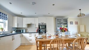 A modern kitchen with white cabinetry, a dining area with a wooden table and chairs, and a kitchen island with a sink and microwave. - 5 Bed in Amroth (oc-pw9068) (Amroth)