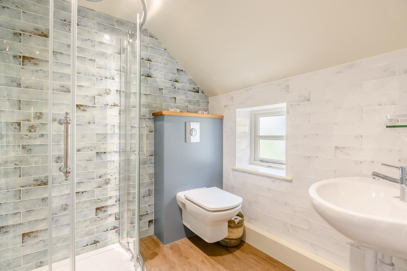 Et badeværelse med brusekabine i glas, toilet, vask og et vindue, der giver naturligt lys.