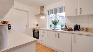 Eine moderne Küche mit weißen Schränken, einem eingebauten Ofen, einem Fenster mit gestreifter Jalousie und einer Arbeitsplatte mit verschiedenen Gegenständen, darunter eine Kaffeemaschine und eine Flasche.