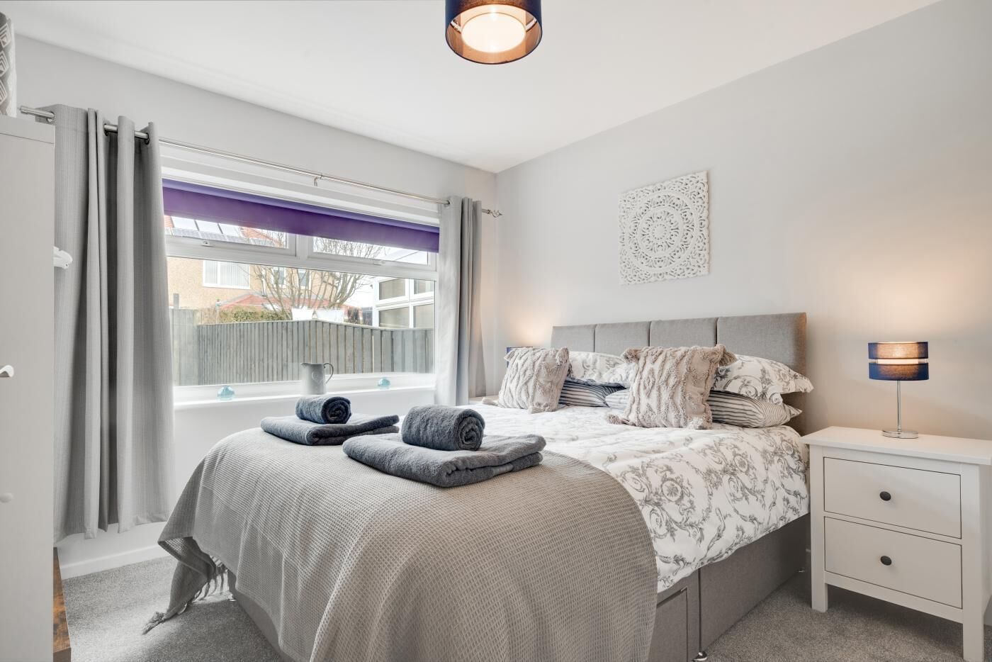 Une chambre à coucher comprenant un lit, une table de chevet, une lampe, une fenêtre avec des rideaux et un élément décoratif mural.
