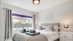 Ein Schlafzimmer mit Bett, Nachttisch, Lampe, Fenster mit Vorhängen und einer Wanddekoration.