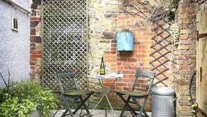 A small outdoor seating area with two metal chairs and a table, a bottle of wine, and a blue barrel against a brick wall. - 2 Bed in Snape (oc-sfbox) (Snape)