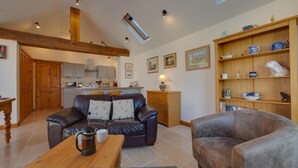 Un salon chaleureux avec un canapé en cuir, une table basse en bois et un fauteuil gris. On aperçoit une bibliothèque en bois ornée d’objets décoratifs, ainsi qu’une cuisine au fond de la pièce.