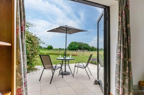 Een open schuifdeur die uitkomt op een terras met een tafel en stoelen, een parasol en uitzicht op een grasveld en bomen.